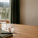 Glaskaraffe, Wasserglas und Notizbuch vor einem Fenster mit Blick auf bewaldete Hügel